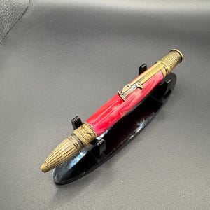 May include: A handcrafted pen with a red and white marbled barrel and brass accents. The pen features a detailed design on the brass components and is displayed on a black pen stand. The pen's tip is a light beige color.
