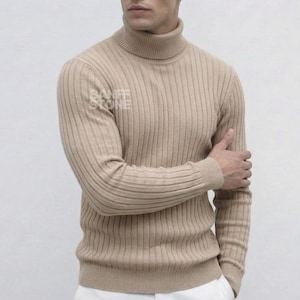 Op de afbeelding: Een beige geribbelde coltrui. De trui heeft een hoge halslijn en lange mouwen. Het model draagt een witte broek. De merknaam "BANFF STONE" is zichtbaar onderaan de afbeelding.