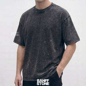 Op de afbeelding: Een donkergrijs, zuur gewassen t-shirt met korte mouwen en een ronde hals. Het shirt heeft een iets oversized pasvorm en een getextureerde uitstraling. De tekst "BANFF STONE" is zichtbaar op het shirt. Het shirt is gemaakt van een zacht, comfortabel materiaal.