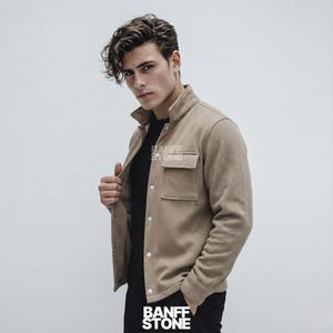 Op de afbeelding: Een beige jasje met twee voorzakken en knopen. Het jasje wordt over een zwart shirt gedragen. De tekst "BANFF STONE" is zichtbaar op het jasje en onderaan de afbeelding.