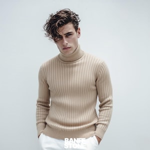 May include: A tan ribbed turtleneck sweater with long sleeves. The sweater is paired with white pants. The brand name "BANFF STONE" is visible at the bottom of the image. The sweater is a neutral color and has a classic design.