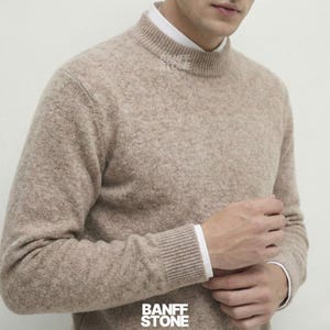 Puede incluir: Un suéter de cuello alto de color beige con mangas largas. El suéter tiene cuello y puños acanalados. Se ve una camisa blanca con cuello debajo. La marca "BANFF STONE" está impresa en la parte inferior de la imagen.