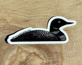 May include: A black and white sticker of a loon, a type of diving bird, with a red eye. The sticker is on a wooden surface. The text "WHITEPINEARTWORKS.COM" is printed on the sticker.