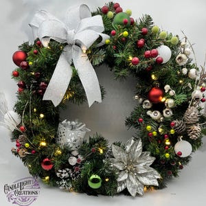 Pode incluir: Uma coroa de Natal prateada e verde com um grande laço prateado, enfeites vermelhos e brancos e uma poinsétia prateada. A coroa é decorada com ramos de pinheiro, bagas e pinhas. A coroa é iluminada com luzes brancas.