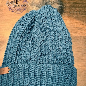 Puede incluir: Un gorro de punto de color azul turquesa con un patrón de trenza y un pompón de piel sintética en la parte superior. El gorro tiene una etiqueta de cuero marrón unida al borde.