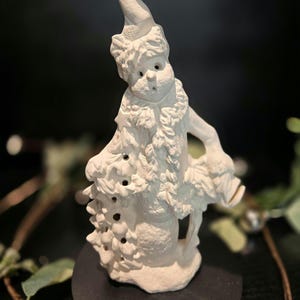 May include: White ceramic figurine of a snowman wearing a hat and holding a small tree. The snowman has a round body and a small face with eyes and a mouth. The figurine is on a black base.
