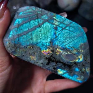 Puede incluir: Una piedra preciosa de labradorita pulida con destellos iridiscentes azules, turquesas y amarillos. La piedra tiene un color base gris oscuro con grietas visibles y una forma suave y redondeada. Otras piedras son visibles en el fondo.