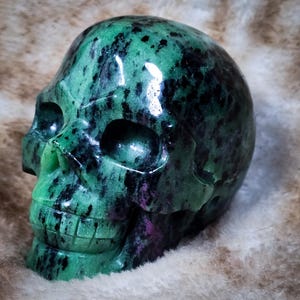 May include: A green and black skull carved from a green gemstone. The skull has a detailed face with a prominent nose and teeth.