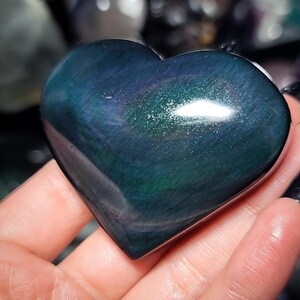May include: A heart-shaped, polished gemstone with a dark teal and green iridescent sheen. The stone has a smooth, reflective surface, and the heart shape is clearly defined. The gemstone is held in a hand, with other objects blurred in the background.