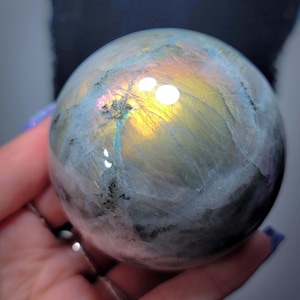 May include: A polished labradorite sphere exhibiting iridescence. The gemstone shows flashes of gold, blue, and green. The sphere is smooth and round, reflecting light and creating a shimmering effect. The colour palette is a mix of cool blues and warm golds.