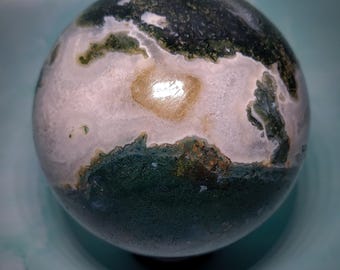 May include: A polished, spherical gemstone with a unique pattern. The orb features a mix of white, green, and tan colours, creating a marbled effect. The stone's surface is smooth and reflective, with a natural, earthy aesthetic. The sphere is a decorative item.