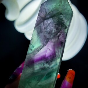 May include: A polished fluorite crystal tower featuring a translucent blend of deep purple and vibrant green hues. The mineral exhibits natural internal fractures and color zoning throughout its elongated, multi-faceted hexagonal prism structure.