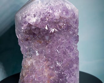 May include: A large amethyst crystal geode with a polished, angled top. The crystal is a deep purple color with a rough, textured surface. The top is a light gray and white banded agate. The geode is displayed on a black surface.