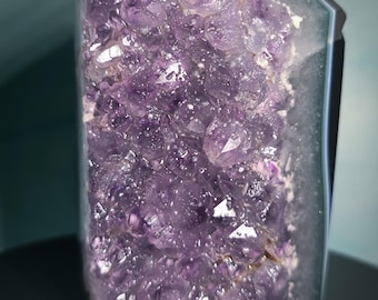 May include: A large, polished amethyst crystal geode. The geode features a deep purple interior with sparkling crystal formations. The exterior is a translucent, light gray color, giving the crystal a striking contrast. The crystal is displayed on a black surface.