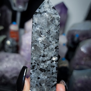 May include: A polished stone tower with a hexagonal point, featuring a speckled pattern of translucent grey and opaque black mineral inclusions throughout the crystalline structure, displaying a smooth, reflective surface with natural variations.