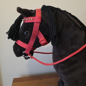 May include: A plush black horse toy featuring a soft dark coat and black yarn mane. The horse is equipped with a bright coral-coloured nylon bridle that includes a decorative row of small clear rhinestones across the browband and a matching coral rope rein.