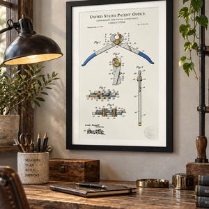 Peut inclure: Une impression de brevet encadrée intitulée United States Patent Office pour un coupe-câble par Louis Rogoff. Le document présente des dessins techniques numérotés de Fig. 1 à Fig. 5, montrant l'outil avec des poignées bleues et des éléments dorés.