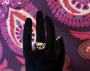May include: A silver-colored ring in the shape of a cat's head is displayed on a black hand. The ring has detailed features, including ears, eyes, and a nose. The background is a purple and pink paisley pattern.