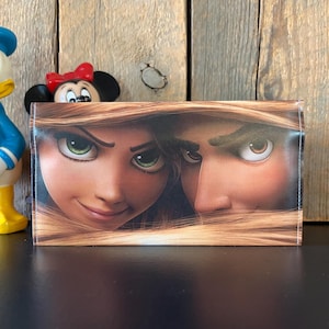 May include: A wallet featuring a close-up of two animated characters with green and brown eyes, framed by long, light brown hair. The wallet is rectangular and appears to be made of a glossy material. A Mickey Mouse figurine and a Donald Duck figurine are in the background.