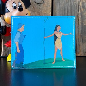 May include: A blue and green embroidered patch featuring a cartoon image of a man and a woman standing on opposite sides of a tree branch. The man is wearing a blue shirt and brown pants. The woman is wearing a brown dress and has long black hair.