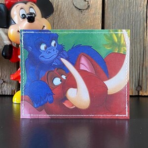 May include: A small, rectangular wallet featuring a colorful illustration of animated characters. The image depicts a blue gorilla and a red warthog with large tusks, set against a green and yellow background. The wallet has white stitching around the edges.