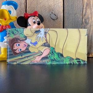 May include: A wallet with a white stitched border features a colorful illustration of a woman in a yellow dress with green foliage. The woman is holding her hand up in the air and has a surprised expression on her face.