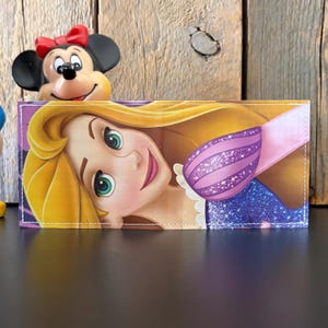May include: A wallet featuring a colorful illustration of a cartoon character with long blonde hair, green eyes, and a pink mouth. The character is wearing a purple and pink dress. A Minnie Mouse figurine is in the background.