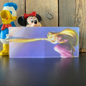 May include: A purple and gold wallet with a picture of a cartoon princess with long blonde hair. The princess is holding a long strand of hair.