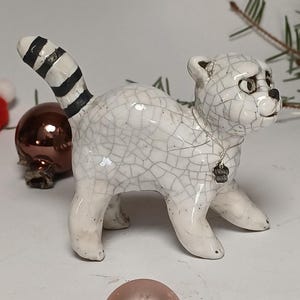 Könnte beinhalten: Eine weiße Keramik-Katzenfigur mit Rissglasur-Effekt. Die Katze hat einen schwarz-weiß gestreiften Schwanz und ein kleines silbernes Etikett mit der Aufschrift "Hand Made". Die Katze steht auf allen Vieren.
