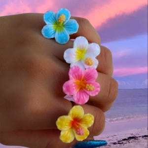 Może przedstawiać: Cztery kolorowe pierścionki z kwiatami w kolorze różowym, białym, żółtym i niebieskim, z motywem hibiskusa. Pierścionki są noszone na palcach osoby.