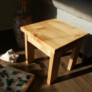 May include: A small, square wooden side table with a natural finish. The table is made of light-colored wood and has four sturdy legs. The table is approximately 18 inches tall and 18 inches wide. A decorative vase and a miniature zen garden are also visible.