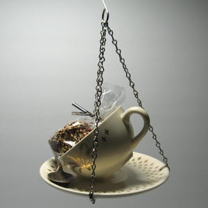 May include: A white teacup and saucer with a gold star pattern hangs from a silver chain. The teacup is filled with birdseed and a clear plastic bag.