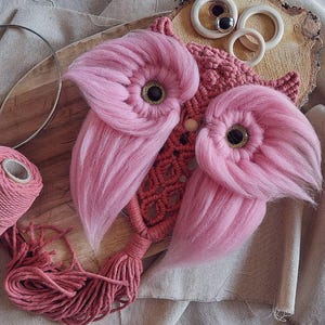 May include: Pink and red yarn owl wall hanging with a wooden base and a tassel. The owl has large pink fluffy wings and black eyes.
