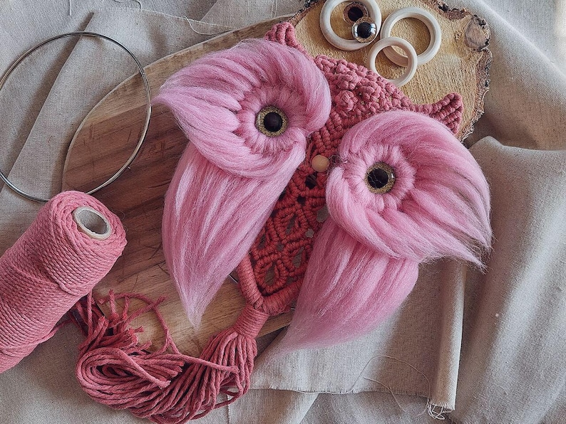 May include: Pink and red yarn owl wall hanging with a wooden base and a tassel. The owl has large pink fluffy wings and black eyes.