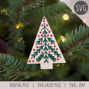 May include: Wooden Christmas tree ornament with green embroidered branches and red berry accents. The ornament is hanging from a green string against a blurred background of a Christmas tree. The text "DIGITAL FILE | SVG LASEF FILE | SVG - DXF" is at the bottom.