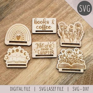 Pode incluir: Cinco placas de madeira cortadas a laser com ditados sobre livros. As placas apresentam um arco-íris, um livro com luas, um livro com flores, um livro com um design floral e o texto "Just one more chapter". As placas são perfeitas para adicionar um toque de fantasia à decoração da sua casa.