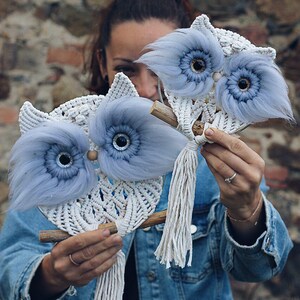 May include: Two blue and white macrame owl wall hangings with fluffy blue feathers around the eyes. The owls are hanging from wooden sticks.