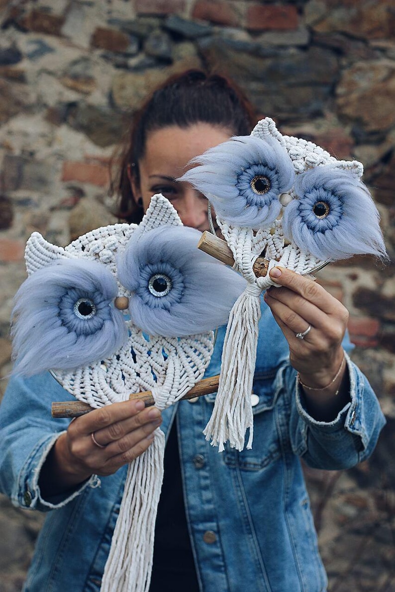 May include: Two blue and white macrame owl wall hangings with fluffy blue feathers around the eyes. The owls are hanging from wooden sticks.