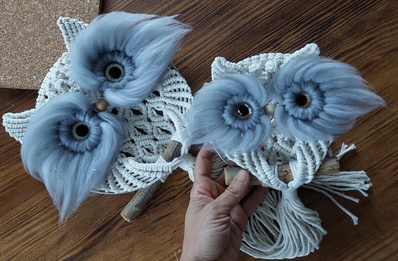 May include: Two macrame owl wall hangings with blue yarn accents. The owls have large, round eyes and are made with white and blue yarn.