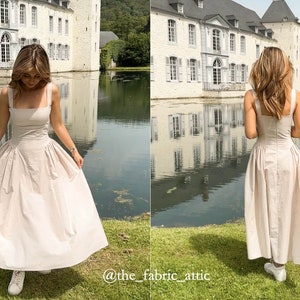 May include: A woman wearing a light beige sleeveless dress with a square neckline and a gathered skirt. She is standing in front of a white building with a pond in the foreground. The text "@the_fabric_attic" is visible in the bottom right corner of the image.