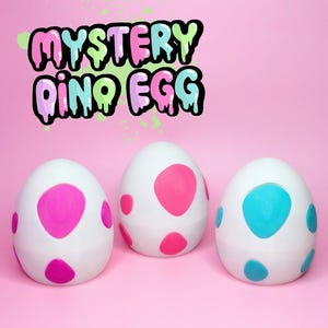 May include: Three white plastic dinosaur eggs with colourful polka dots. The eggs are pink, blue, and pink. The text "MYSTERY DINO EGG" is written in colourful letters above the eggs.
