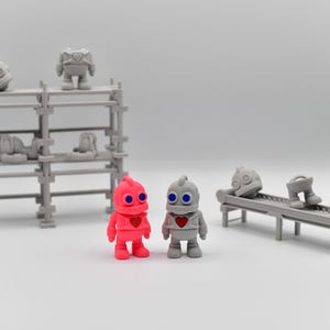 Könnte beinhalten: Eine skurrile Szene mit Miniatur-Roboterfiguren und Regalen im Industriestil. Zwei Roboter stehen im Vordergrund, einer rosa und einer grauer, jeder mit einem Herz auf der Brust. Weitere Roboterfiguren befinden sich auf Regalen und einem Förderband.