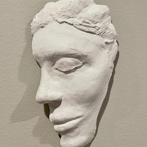 Puede incluir: Escultura de yeso blanco de un rostro humano, vista de perfil, con los ojos cerrados y una leve sonrisa.