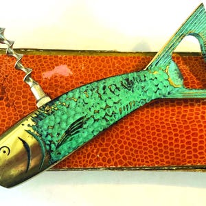 May include: A vintage fish-shaped bottle opener and corkscrew. The opener is made of brass and turquoise-colored metal, with a corkscrew on top. It rests on a textured, orange, rectangular box.