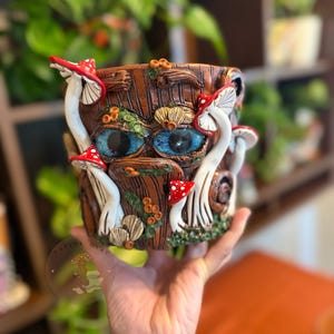 Op de afbeelding: Een bruine cilindrische plantenbak met een textuur die op schors lijkt, voorzien van twee grote, felblauwe ogen en diverse gebeeldhouwde rood-witte paddenstoelen. De buitenkant is versierd met kleine groene accenten en bruine houtdetails.