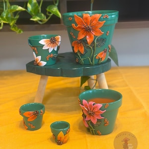 Puede incluir: Una colección de cinco macetas de terracota verdes de varios tamaños, cada una decorada con apliques florales de arcilla tridimensionales en naranja o rosa. Las macetas presentan pétalos esculpidos, hojas y pequeños acentos circulares. Un logotipo es visible.