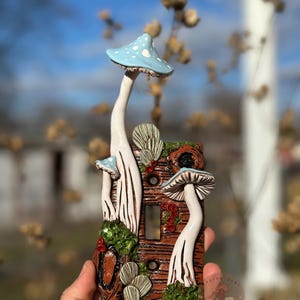 May include: A decorative light switch cover featuring a whimsical mushroom design. The cover is brown with white and blue mushrooms, green moss, and red berries. The mushrooms have white stems and blue caps with white polka dots. The design is hand-painted.