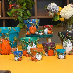 May include: A collection of various sized plant pots in colors including teal, blue, green, orange, purple, and black. Each pot features three-dimensional sculpted orange and white pumpkins, green vines, and small red berries attached to the exterior.