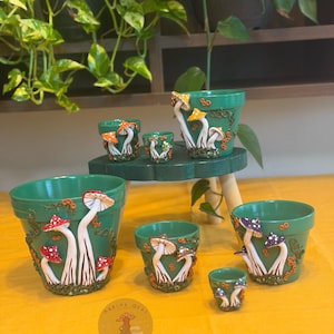 Può includere: Una collezione di sei vasi da fiori in ceramica verde di varie dimensioni, ciascuno decorato con funghi scolpiti in tre dimensioni nei colori rosso, arancione e viola, insieme a dettagli di viti testurizzate. È visibile un logo circolare con il testo Marina Olas.