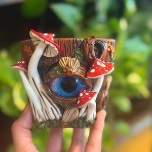 May include: A brown cylindrical planter with a textured bark-like surface featuring a large, realistic blue eye in the center. The planter is decorated with several white and red-spotted mushrooms and small brown leaf-like accents.